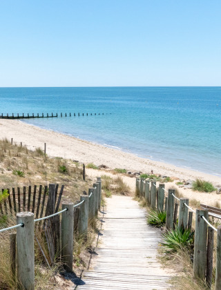 Træsti fører til en rolig sandstrand ved L'Aiguillon-sur-Mer, omgivet af klitter og blåt hav.