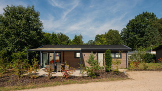 Vue extérieure de la Veluwe Lodge par une journée ensoleillée, entourée d’arbres, de buissons et d’une terrasse.