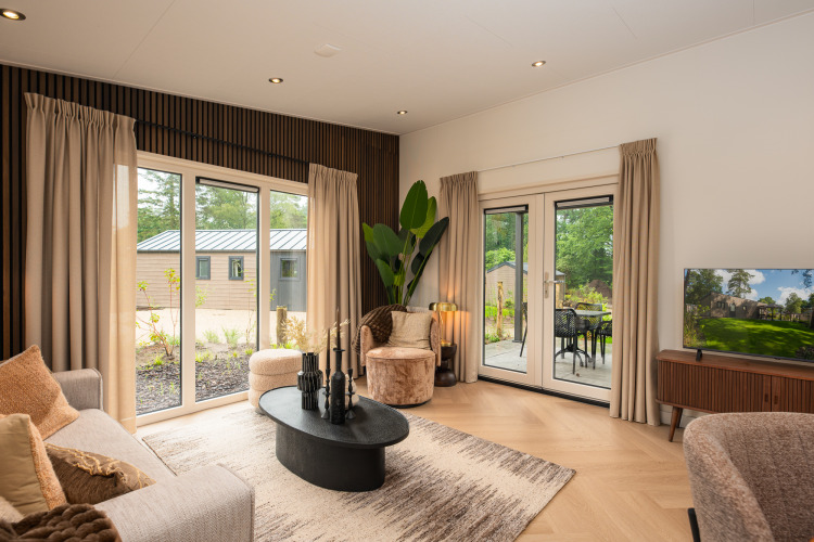 Modernes Wohnzimmer in einer Lodge mit großen Fenstern, beigen Vorhängen und Ausblick ins Grüne.