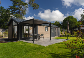 Alloggio glamping moderno con patio, sedie da esterno e prato verde circondato da alberi alti.