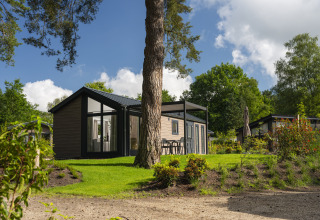 Moderne lodge met grote ramen en terras, omringd door groen en bomen onder een blauwe hemel.