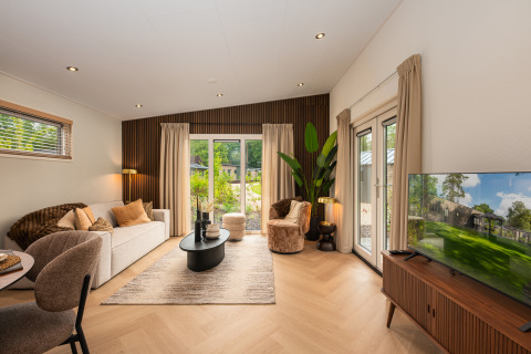 Moderne lodge-stue med træpaneler, store vinduer, elegante møbler og udsigt til naturen udenfor.