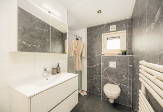 Moderne lodgebadeværelse med grå marmorgulve, væghængt toilet, hvid vask og en badekåbe ved bruseren.