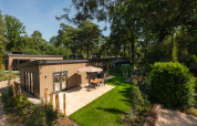 Modern Loevestein lodge with hot tub, sunlit patio, lush green garden, surrounded by tall trees.