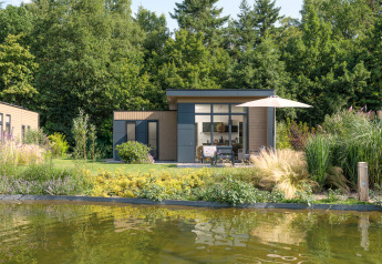 Moderne lodge med store vinduer, terrasse og parasol ved en dam i grønne omgivelser på Veluwepark de Bosgraaf.