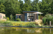 Moderne lodge met grote ramen, terras en parasol aan een vijver in Veluwepark de Bosgraaf, Nederland.