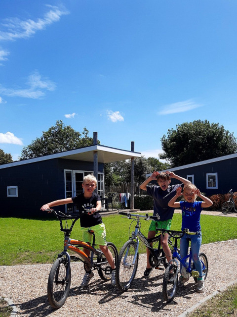 Drie kinderen poseren met hun fietsen voor moderne chalets op een zonnige dag bij Kompas Camping Nieuwpoort.