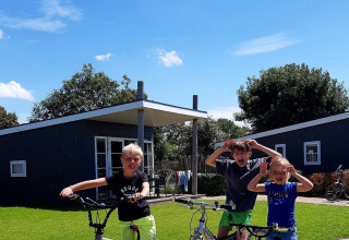 Drie kinderen poseren met hun fietsen voor moderne chalets op een zonnige dag bij Kompas Camping Nieuwpoort.