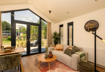 Lyst tiny house-stue med store vinduer, sofa, spisebord og moderne indretning med udsigt til haven.