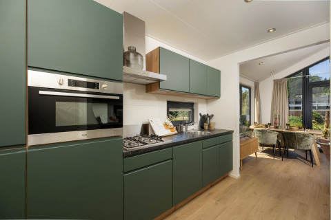 Cuisine moderne de tiny house avec placards verts, coin repas, grandes fenêtres et vue sur le jardin.