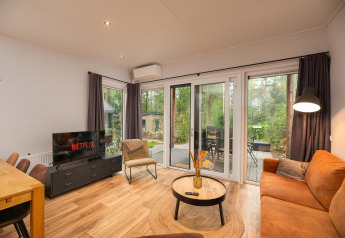 Living room with sofa, TV displaying Netflix, dining table and large windows at Sand Lodge Comfort, Veluwepark de Bosgraaf.