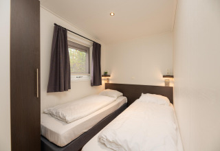 Moderne lodgeværelse med to enkeltsenge, hvidt sengetøj, vindue og mørke gardiner i Veluwepark de Bosgraaf.