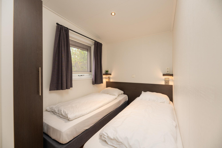 Modern lodge bedroom with two single beds, white bedding, window and dark curtains at Veluwepark de Bosgraaf.