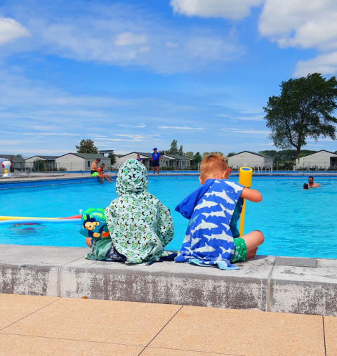 Twee kinderen ontspannen bij het zwembad van Kompas Camping Nieuwpoort, onder een blauwe lucht.
