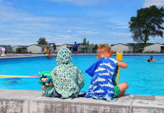 Twee kinderen ontspannen bij het zwembad van Kompas Camping Nieuwpoort, onder een blauwe lucht.