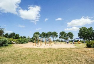 Zicht op een speelplein met schommels en klimrekken, omgeven door groen op Kompas Camping Nieuwpoort.