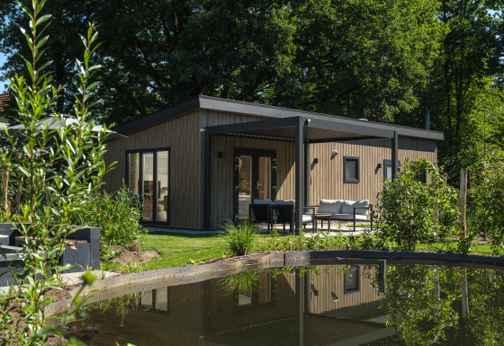 Moderne Loevestein Deluxe-lodge med hot tub, omgivet af natur, terrasse og lille dam i forgrunden.