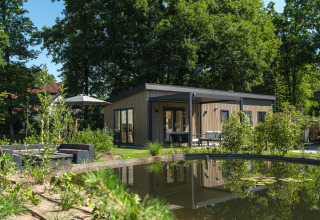 Moderne Loevestein Deluxe-hytte med spabad ved dammen i Veluwepark de Bosgraaf, Holland, i grønne omgivelser.