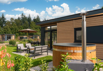 Outdoor area of Lodge Cannenburch featuring a hot tub, garden furniture, and lush greenery under blue sky.