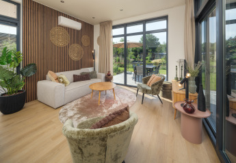 Moderne stue i en lodge på Veluwepark de Bosgraaf med store vinduer, udsigt til terrasse og lyse møbler.