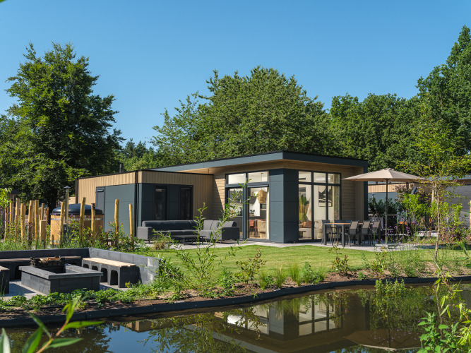 Moderne Essenburgh Deluxe Lodge mit Whirlpool, Terrasse, Gartenmöbeln und Teich, umgeben von Natur.