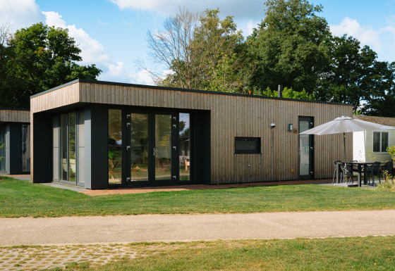 Moderne Happy Oak Lodge met grote ramen, houten gevel en tuinset met parasol in groene omgeving.