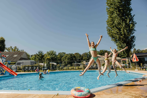 Tre glade børn hopper i poolen på en glampingplads, mens andre børn leger i vandet under en klar himmel.
