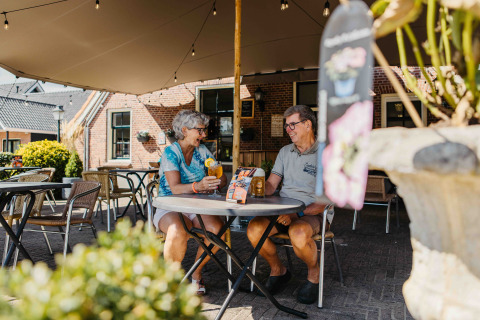 Twee oudere mensen genieten van drankjes en een babbel aan een tafel bij een glampingverblijf.