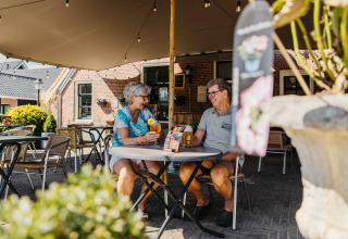 Twee oudere mensen genieten van drankjes en een babbel aan een tafel bij een glampingverblijf.