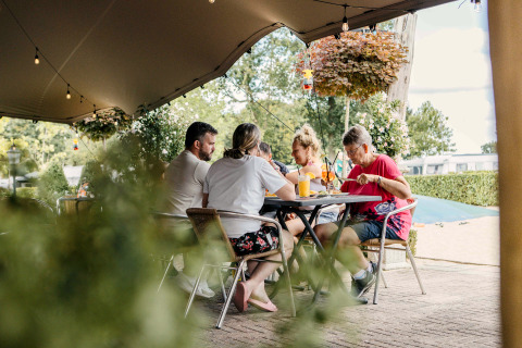 A family enjoys food and drinks at a table under a canopy, relaxing at a glamping site with greenery.