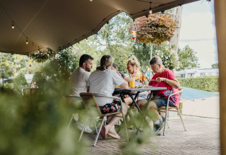 En familie nyder mad og drikke under et telt på et glampingophold med grøn natur i baggrunden.