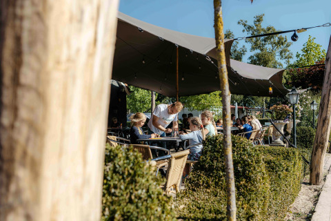 Outdoor café seating at a glamping site with people dining under a canopy on a bright sunny day.