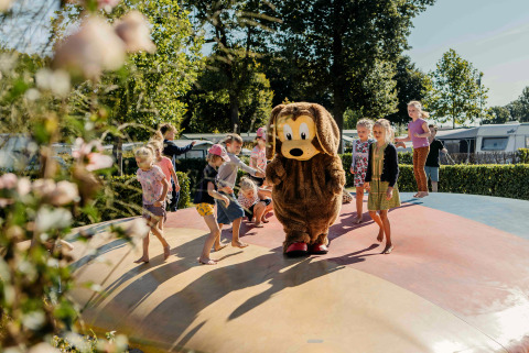 Children play on an inflatable pillow at a glamping site, joined by a person in a dog costume on a sunny day.