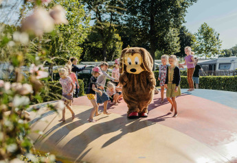 Children play on an inflatable pillow at a glamping site, joined by a person in a dog costume on a sunny day.