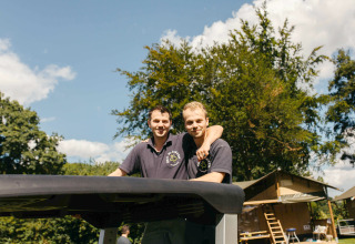 Two men in dark polo shirts smiling together outside at a glamping accommodation on a sunny day.