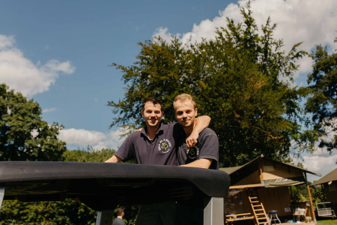To mænd iført sorte poloshirts står smilende sammen udendørs ved glamping-overnatning på en solrig dag.