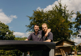 Twee mannen in zwarte polo’s glimlachen samen buiten aan een glamping-verblijf op een zonnige dag.