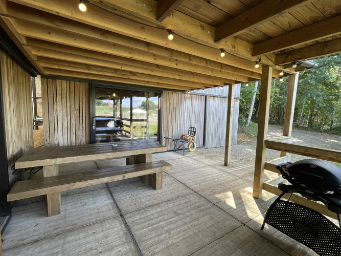 Covered terrace with a wooden picnic table, benches, and a grill at a tiny house in Limburg forest, Belgium.