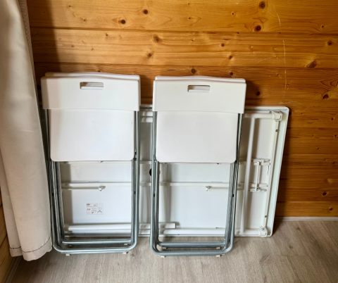Twee opvouwbare stoelen tegen een houten wand in een hut, met een opgevouwen tafel erachter.