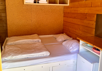 Innenansicht einer Basic-Blockhütte mit Doppelbett, Bettwäsche, Regal, Handtüchern und Minikühlschrank.