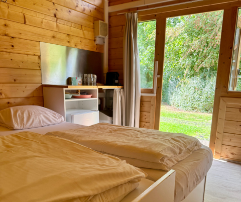 Interieur van een eenvoudige blokhut met tweepersoonsbed, houten muren en tuinzicht in Ieper, België.