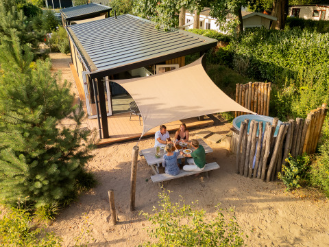 Fire personer nyder udendørs frokost ved et picnicbord foran Sand Lodge Jacuzzi tiny house med solsejl.