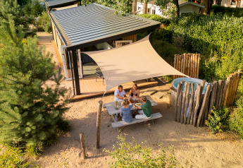 Quatre personnes partagent un repas dehors devant le tiny house Sand Lodge Jacuzzi couvert d’une voile d’ombrage.