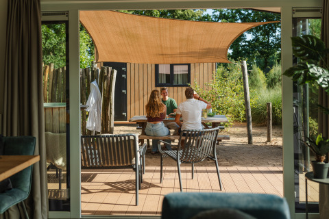 Udendørs scene ved et lille hus, hvor tre personer sidder ved et bord under et solsejl i Sand Lodge Jacuzzi.