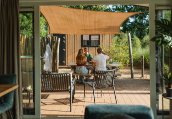 Udendørs scene ved et lille hus, hvor tre personer sidder ved et bord under et solsejl i Sand Lodge Jacuzzi.