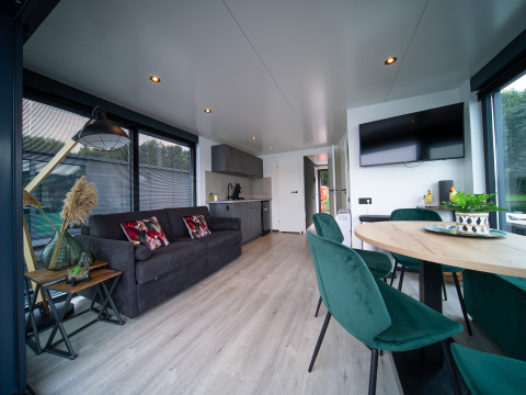 Moderne glampingaccommodatie met leefruimte, keukenhoek, zetel, eettafel en groene stoelen bij ramen.