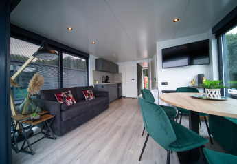 Modern glamping accommodation with living area, kitchenette, sofa, dining table, and green chairs by windows.