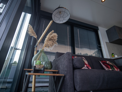 Modern glamping-interieur met grijze bank, kussens, vloerlamp, planten en grote ramen met jaloezieën.