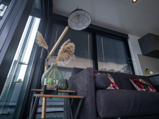 Modernes Glamping-Interieur mit grauem Sofa, Beistelltisch, Pflanzen und großen Fenstern mit Jalousien.