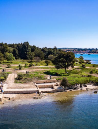 Kystlandskab nær Medulin, Istrien, Kroatien, med gamle ruiner, grønne træer og blå havvand under klar himmel.